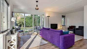 Una sala de estar moderna con un sofá morado, un televisor de pantalla plana y un comedor con una mesa y sillas blancas.