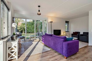Un salon moderne avec un canapé violet, un téléviseur à écran plat et un coin repas comprenant une table et des chaises blanches.