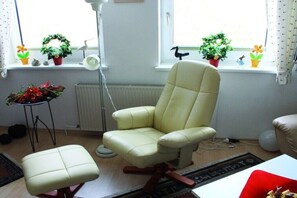 A room with a cream-colored armchair, a matching ottoman, a floor lamp, and a radiator. There are potted plants on the windowsill and a small table with a plant. - Holiday apartment with 2 bedrooms (Grünendeich)