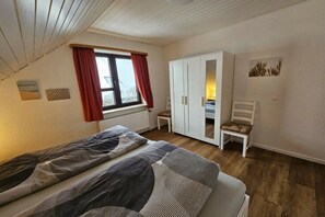 A bedroom with a bed, a wardrobe, a chair, and a window with curtains. - Holiday apartment 3 House Froschblick (Greetsiel)