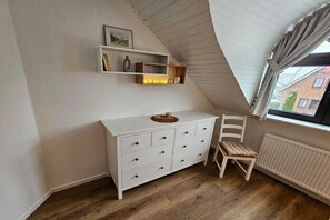 Une pièce avec un plafond en pente, une commode blanche à tiroirs, une chaise et une fenêtre avec des rideaux.