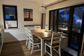 Un coin repas avec une table en bois et des chaises blanches, un luminaire suspendu et une fenêtre donnant sur des immeubles.