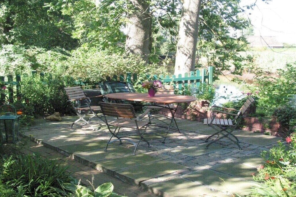 An outdoor patio with a round table and four chairs, surrounded by lush greenery and a brick wall.