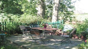 En udendørs terrasse med et rundt bord og fire stole, omgivet af frodig beplantning og en murstensmur.