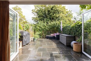 Ein überdachter Patio-Bereich mit Sitzgelegenheit, Grill und Topfpflanzen.