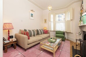 Una sala de estar con un sofá color beige, una mesa de centro de vidrio y un sillón verde. Hay una chimenea con repisa y una ventana con persianas blancas.