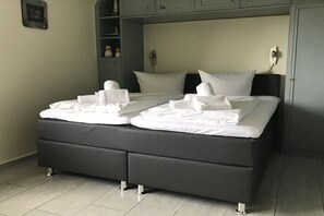 Una cama doble con sábanas y toallas blancas, un cabecero tapizado en gris y un estante con adornos.