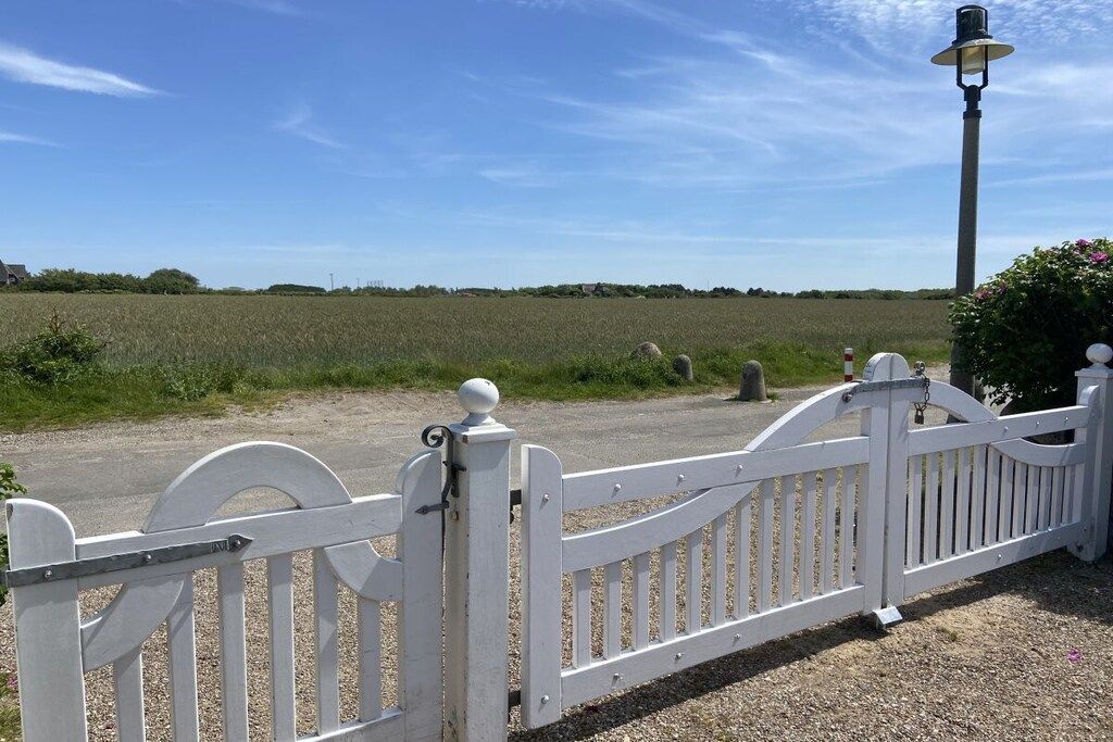 A white metal gate with a curved top, a lamp post, and a gravel path leading into a field.