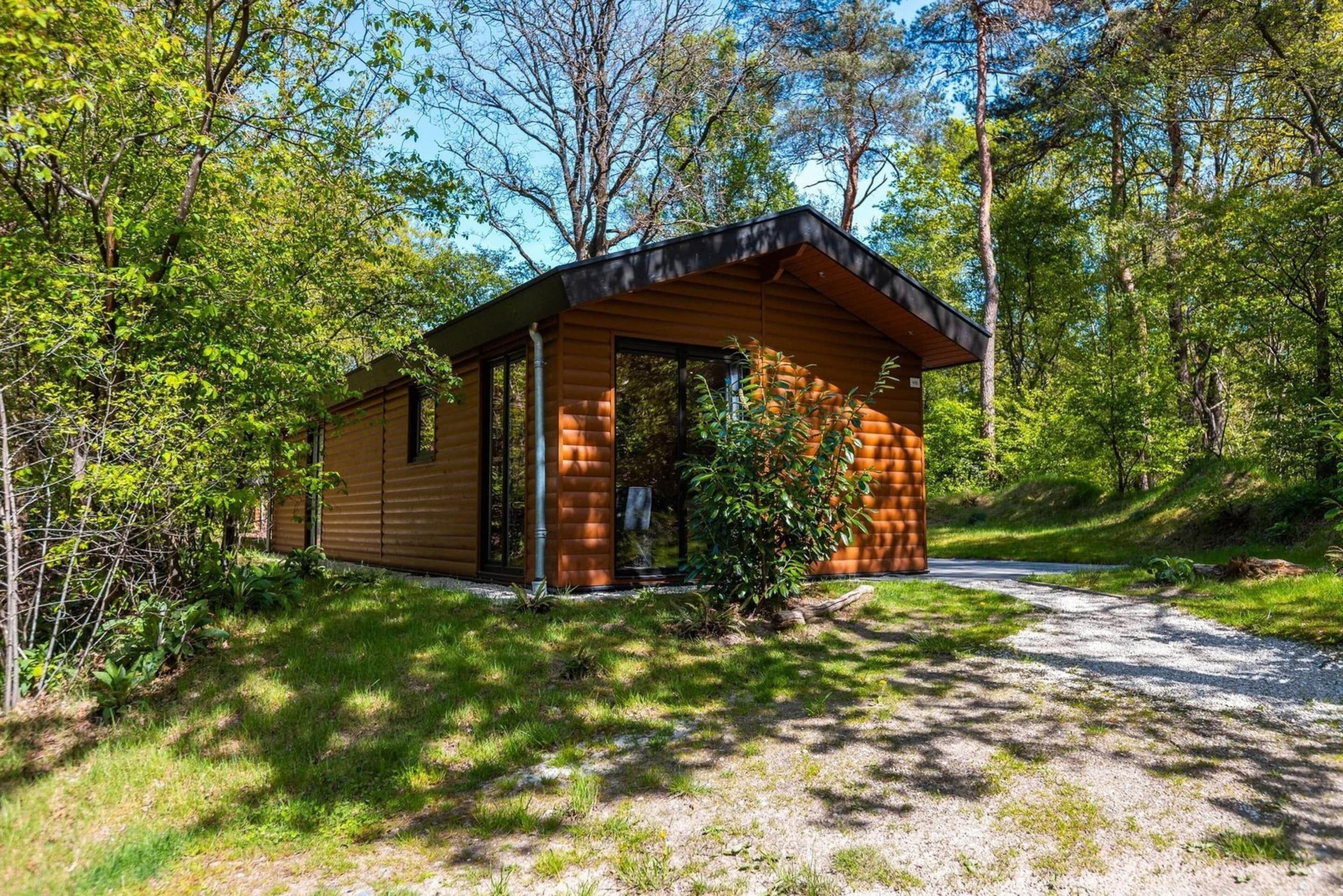 Eine Hütte mit geneigtem Dach, großen Fenstern und einer Tür, umgeben von Bäumen und einem Kiesweg.