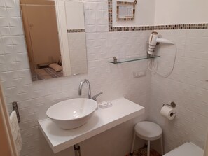 Une salle de bain avec un lavabo blanc, un miroir, des toilettes et une étagère en verre où sont rangés les articles de toilette.