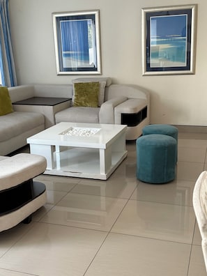 Uma sala de estar moderna com um sofá branco, uma mesa de centro branca e um banquinho verde-água. Há duas imagens emolduradas na parede.