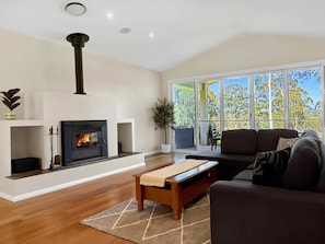 A modern living room with a fireplace, a sofa, a coffee table, and a view through large windows. - North Peak Manor - Spacious home with room for everyone (Pokolbin)