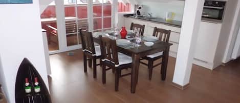 Comedor con una mesa dispuesta para cuatro, cocina al fondo y vista a través de un ventanal.