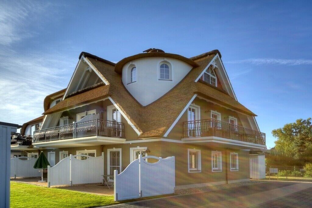 Une maison à deux étages dotée d’un toit triangulaire au design unique, d’un balcon à l’étage supérieur et d’une allée d’accès fermée par un portail.