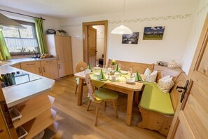 Ein gemütlicher Essbereich mit einem Holztisch, gedeckt für zwei Personen, einer Bank mit Kissen und Blick ins Freie durch ein Fenster.