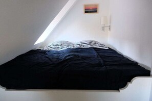 Ein Einzelbett in einem Zimmer mit schrägem Dach, ein kleines Bild an der Wand und eine Wandleuchte.