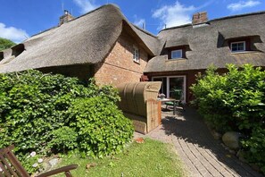 A traditional thatched-roof house with a brick exterior, a paved pathway, and a garden area with a wicker chair and greenery. - Hahnenhof Föhr Apartment 9 (Wrixum)