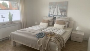 Een slaapkamer met een bed, kussens, een nachtkastje, een lamp, een raam met jaloezieën, een plant en een schilderij van zeilboten.