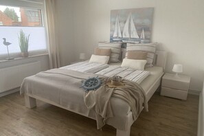 A bedroom with a bed, pillows, a nightstand, a lamp, a window with blinds, a plant, and a painting of sailboats. - Holiday apartment with 1 bedroom (Maasholm)