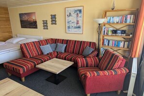 Ein gemütliches Wohnzimmer mit einem gestreiften Sofa, einem kleinen Couchtisch, einem Bücherregal und einem Bett mit weißem Kopfteil.