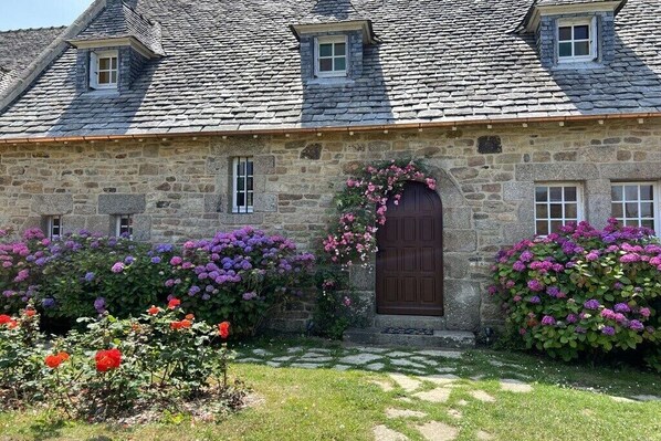 Un bâtiment en pierre avec une porte brun foncé, entouré d’un jardin où poussent des fleurs violettes et rouges.