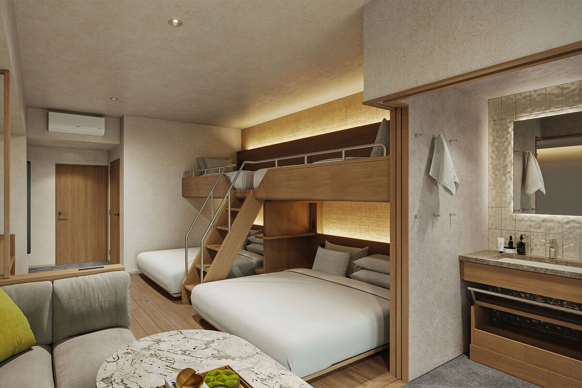 Ein kompaktes Hotelzimmer mit Etagenbett, Schlafsofa, kleinem Tisch und einem Badezimmer mit Spiegel und Waschbecken.