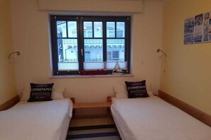 Ein Zimmer mit zwei Betten, einem Fenster mit Blick auf Gebäude und einer kleinen Vitrine mit einem Modellschiff.