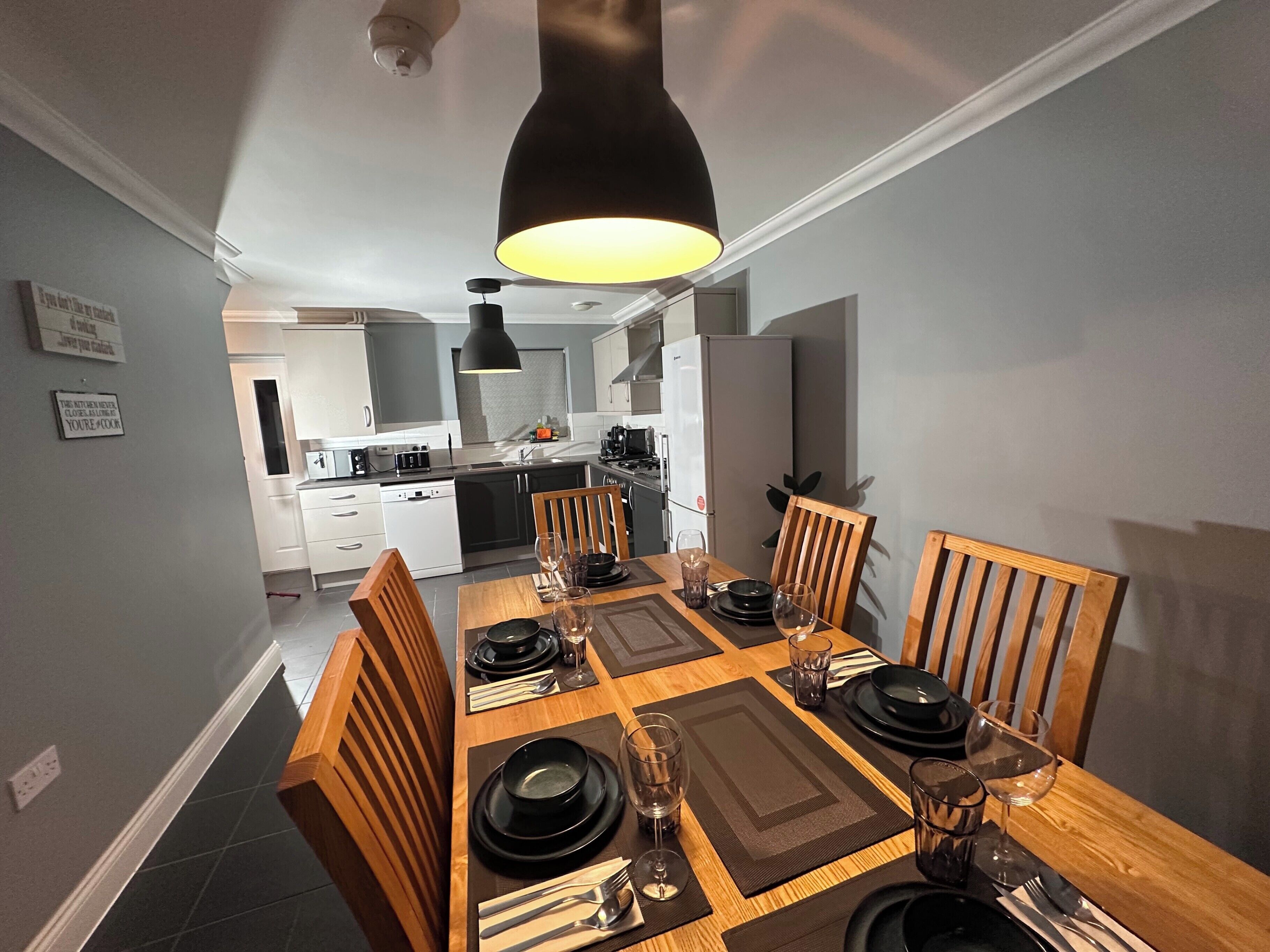 A dining area with a wooden table set for four, surrounded by chairs, in a modern kitchen with white cabinets and a black pendant light.