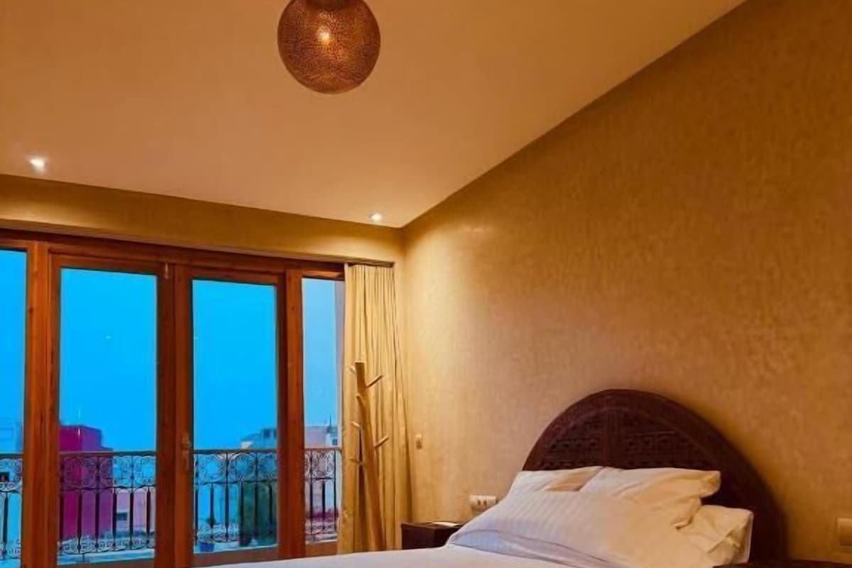 A hotel room with a large bed, a nightstand, a hanging lamp, and a view of the city through large windows.