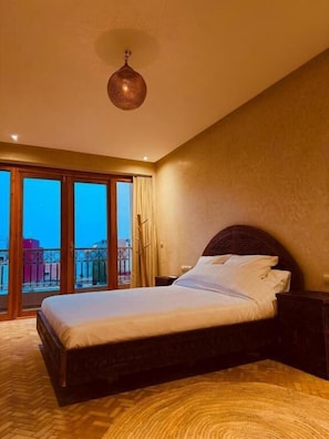 A hotel room with a large bed, a nightstand, a hanging lamp, and a view of the city through large windows.