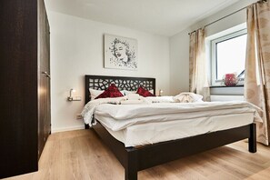 A bedroom with a bed, a headboard, a wall-mounted light, a window with curtains, and a wall art piece. - Holiday apartment in Hörnum with Wi-Fi (Hörnum)