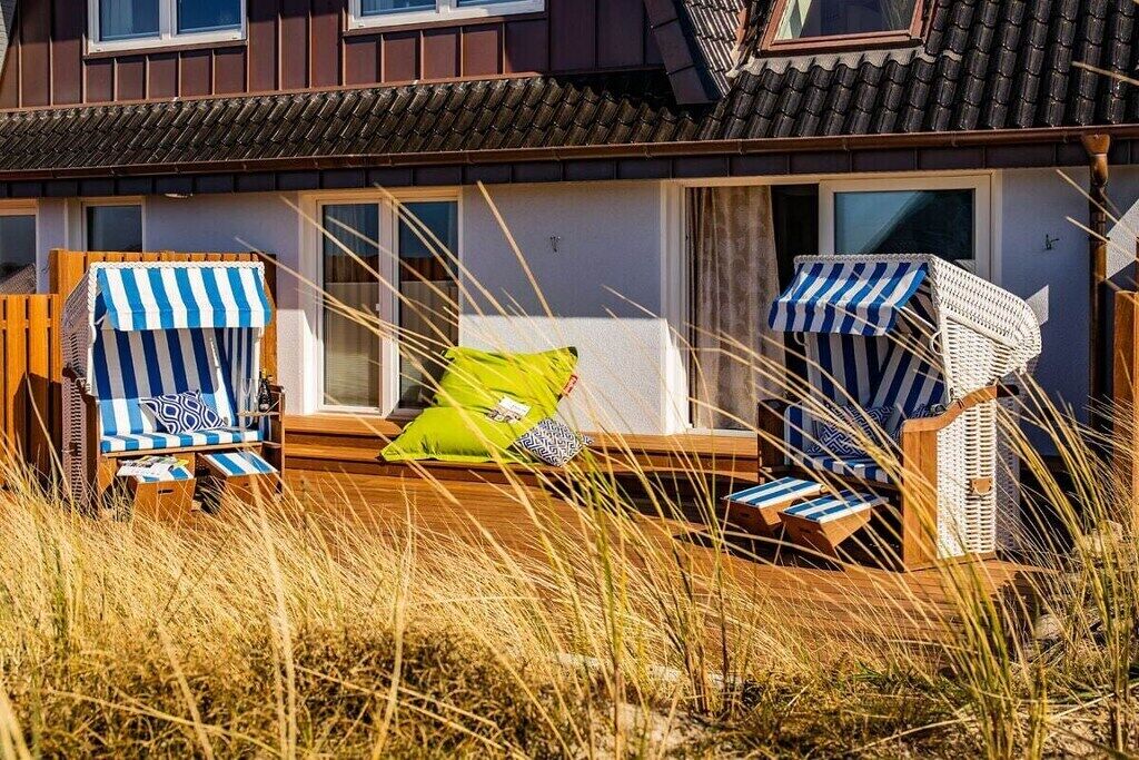 Een pand aan het strand met een houten terras, twee strandstoelen met streepjeszonnedoeken en een enkele ligstoel met kussen.