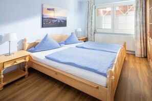 A double bed with a wooden frame and blue bedding, a bedside table with a lamp, a window with curtains, and a painting on the wall. - Holiday apartment in Juist with swimming pool (Juist)