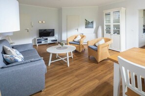 A living room with a sofa, two wicker chairs, a small round table, a television, and a cabinet with glass doors. - Holiday apartment in Juist with swimming pool (Juist)