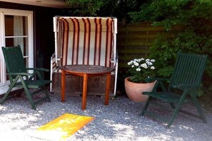 Eine Terrasse mit einem runden Holztisch, zwei grünen Stühlen, einem streifenmusterbezogenen Sitzkissen und einem Topf mit weißen Blüten.