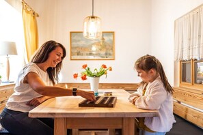 Eine Frau und ein Kind sitzen an einem Holztisch mit einem Schachbrett in einem Raum, der eine Pendelleuchte und ein gerahmtes Bild an der Wand hat.