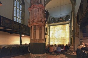 An interior with a large illuminated stained glass window, a central ornate column, and a group of people seated.