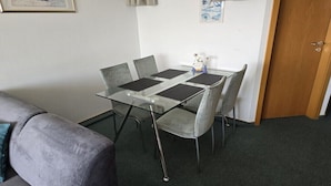 Een kleine kamer met een tafel met glazen blad en vier grijze stoelen, een grijze bank en een houten deur.