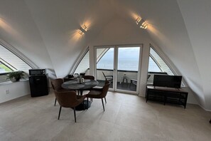 Een eetruimte met een tafel, stoelen, een televisie en uitzicht op zee door grote ramen.