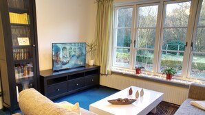 Ein Wohnzimmer mit Fernseher, Sofa, Couchtisch und einem Fenster mit Pflanzen.