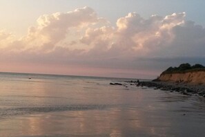 Ein Strand mit ruhiger See, sandigem Ufer und einer Klippe mit Vegetation.