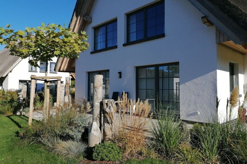 Une maison moderne à deux étages, avec une façade blanche, de grandes fenêtres et un jardin bien entretenu où poussent différentes plantes et un arbre.