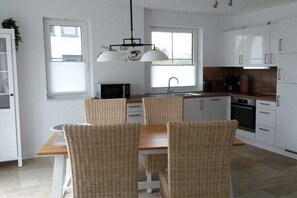 Une cuisine moderne avec des meubles blancs, une table à manger entourée de chaises en osier, un four à micro-ondes et un luminaire suspendu.