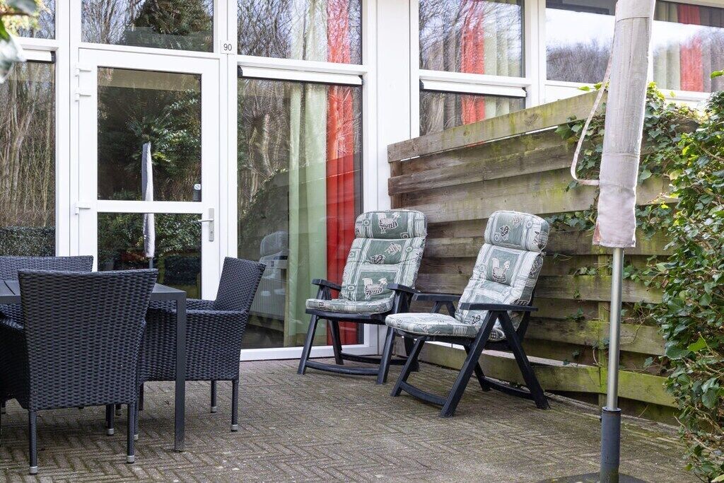 Eine Terrasse mit Korbmöbeln und gemusterten Stühlen, ein Holzzaun und eine Glastür, auf der die Nummer 90 zu sehen ist.