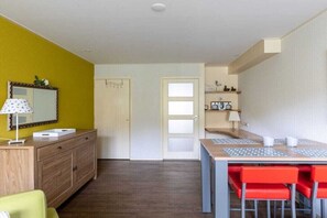 Une pièce de taille modeste, comprenant une commode en bois, un bureau avec des chaises, un miroir et une porte donnant sur une autre pièce.