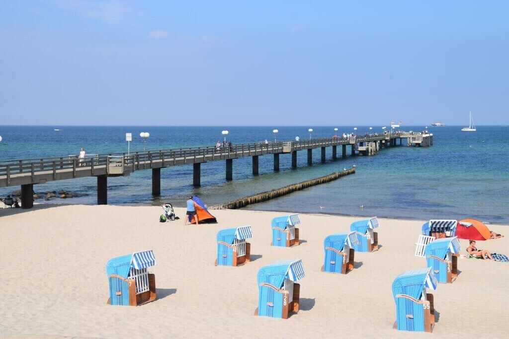 A beach with rows of blue beach chairs and umbrellas, a pier extending into the sea, and a few boats in the distance.