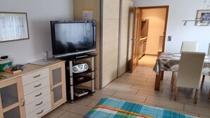 En kompakt opholdsstue med fjernsyn, tekøkken, spiseplads og et farverigt tæppe.