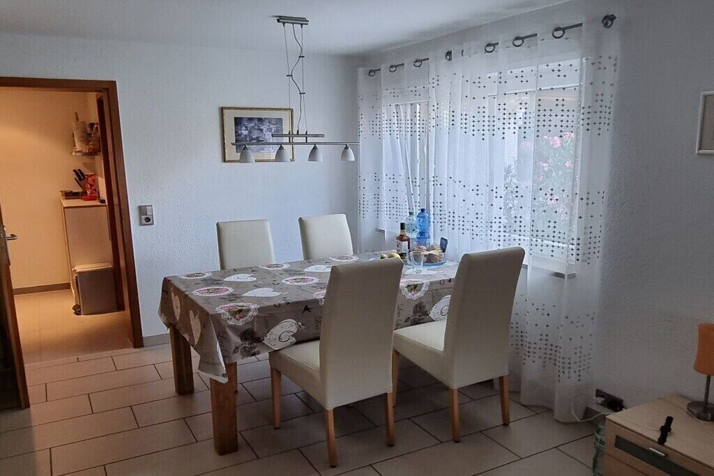 A dining area with a table set for four, chairs, a hanging light fixture, and a window with patterned curtains.