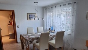 Spiseområde med et bord dækket til fire personer, stole, en hængende lampe og et vindue med mønstrede gardiner.