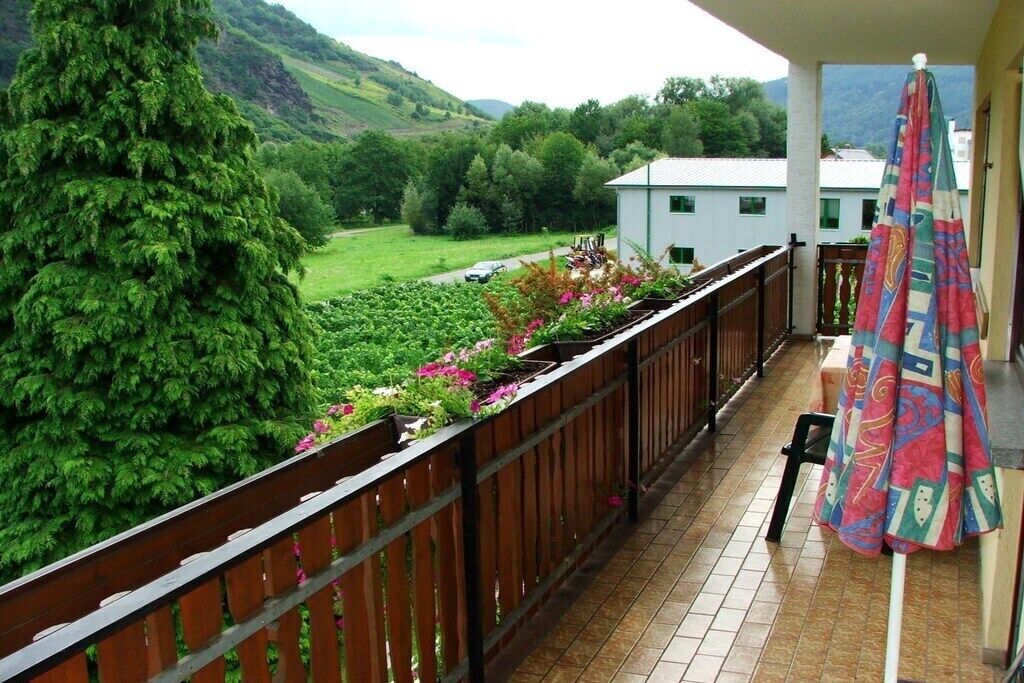 En balkon med udsigt over en frodig have og en hvid bygning i baggrunden.